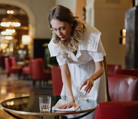 Ce qu’il faut savoir sur l’attaque de la droite contre le salaire minimum Une serveuse en uniforme blanc dessert une table dans un élégant salon d'hôtel ou de restaurant. Elle place un verre d'eau sur une table ronde noire, avec professionnalisme et attention. L'arrière-plan montre un intérieur raffiné avec des fauteuils rouges, un éclairage tamisé et une architecture classique. L'ambiance générale reflète un service hôtelier de qualité dans un établissement luxueux.