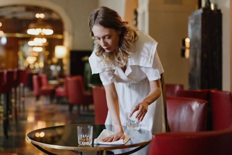 Une serveuse en uniforme blanc dessert une table dans un élégant salon d'hôtel ou de restaurant. Elle place un verre d'eau sur une table ronde noire, avec professionnalisme et attention. L'arrière-plan montre un intérieur raffiné avec des fauteuils rouges, un éclairage tamisé et une architecture classique. L'ambiance générale reflète un service hôtelier de qualité dans un établissement luxueux.