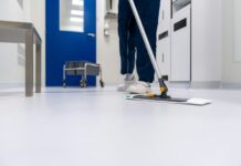 Arrêt du Tribunal fédéral : les salaires minimaux à Zurich et Winterthour sont valides Personne en tenue professionnelle nettoyant un sol blanc brillant avec une vadrouille plate dans un couloir moderne aux murs bleus et blancs. Un chariot de nettoyage est visible en arrière-plan. L'environnement apparaît être un établissement médical ou institutionnel.
