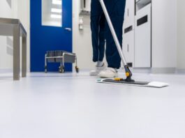 Arrêt du Tribunal fédéral : les salaires minimaux à Zurich et Winterthour sont valides Personne en tenue professionnelle nettoyant un sol blanc brillant avec une vadrouille plate dans un couloir moderne aux murs bleus et blancs. Un chariot de nettoyage est visible en arrière-plan. L'environnement apparaît être un établissement médical ou institutionnel.