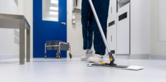 Personne en tenue professionnelle nettoyant un sol blanc brillant avec une vadrouille plate dans un couloir moderne aux murs bleus et blancs. Un chariot de nettoyage est visible en arrière-plan. L'environnement apparaît être un établissement médical ou institutionnel.