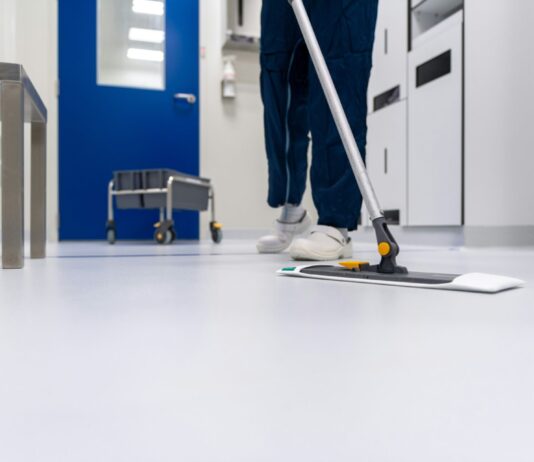 Arrêt du Tribunal fédéral : les salaires minimaux à Zurich et Winterthour sont valides Personne en tenue professionnelle nettoyant un sol blanc brillant avec une vadrouille plate dans un couloir moderne aux murs bleus et blancs. Un chariot de nettoyage est visible en arrière-plan. L'environnement apparaît être un établissement médical ou institutionnel.