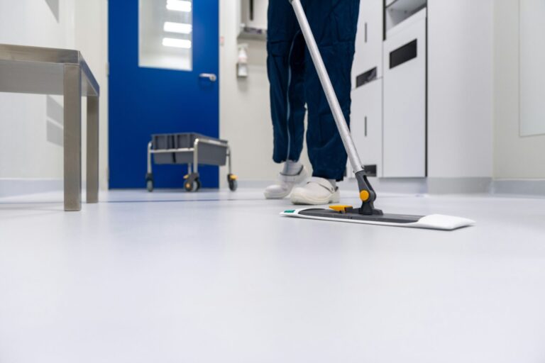 Personne en tenue professionnelle nettoyant un sol blanc brillant avec une vadrouille plate dans un couloir moderne aux murs bleus et blancs. Un chariot de nettoyage est visible en arrière-plan. L'environnement apparaît être un établissement médical ou institutionnel.
