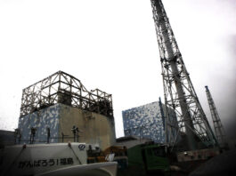 Énergie nucléaire : le risque d’un accident majeur est bien réel Vue de la centrale nucléaire de Fukushima Daiichi montrant deux réacteurs avec leurs structures de confinement en béton recouvertes de panneaux bleus, surplombées de tours de refroidissement métalliques en treillis, sous un ciel gris. Au premier plan, des véhicules de construction et une signalisation en japonais.