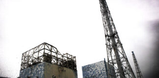 Énergie nucléaire : le risque d’un accident majeur est bien réel Vue de la centrale nucléaire de Fukushima Daiichi montrant deux réacteurs avec leurs structures de confinement en béton recouvertes de panneaux bleus, surplombées de tours de refroidissement métalliques en treillis, sous un ciel gris. Au premier plan, des véhicules de construction et une signalisation en japonais.