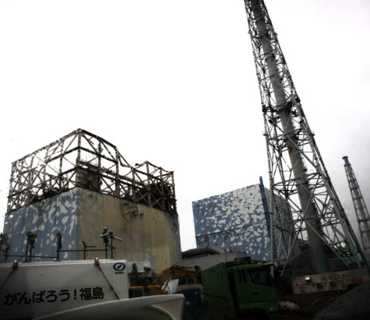 Énergie nucléaire : le risque d’un accident majeur est bien réel Vue de la centrale nucléaire de Fukushima Daiichi montrant deux réacteurs avec leurs structures de confinement en béton recouvertes de panneaux bleus, surplombées de tours de refroidissement métalliques en treillis, sous un ciel gris. Au premier plan, des véhicules de construction et une signalisation en japonais.