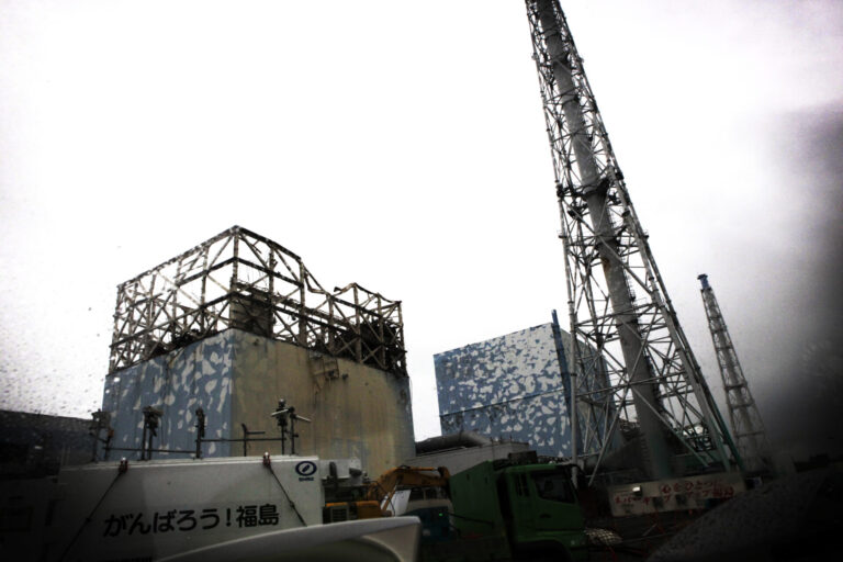 Vue de la centrale nucléaire de Fukushima Daiichi montrant deux réacteurs avec leurs structures de confinement en béton recouvertes de panneaux bleus, surplombées de tours de refroidissement métalliques en treillis, sous un ciel gris. Au premier plan, des véhicules de construction et une signalisation en japonais.