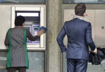 En Suisse, l’écart salarial continue de se creuser Deux personnes se tiennent devant la façade d'une banque. À gauche, une femme portant un gilet rayé vert et blanc nettoye un distributeur automatique de billets (DAB). À droite, un homme en costume bleu marine, tenant une serviette noire, attend devant la porte d'entrée de l'établissement bancaire.