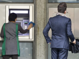 En Suisse, l’écart salarial continue de se creuser Deux personnes se tiennent devant la façade d'une banque. À gauche, une femme portant un gilet rayé vert et blanc nettoye un distributeur automatique de billets (DAB). À droite, un homme en costume bleu marine, tenant une serviette noire, attend devant la porte d'entrée de l'établissement bancaire.