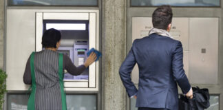 En Suisse, l’écart salarial continue de se creuser Deux personnes se tiennent devant la façade d'une banque. À gauche, une femme portant un gilet rayé vert et blanc nettoye un distributeur automatique de billets (DAB). À droite, un homme en costume bleu marine, tenant une serviette noire, attend devant la porte d'entrée de l'établissement bancaire.