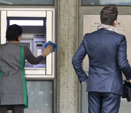 En Suisse, l’écart salarial continue de se creuser Deux personnes se tiennent devant la façade d'une banque. À gauche, une femme portant un gilet rayé vert et blanc nettoye un distributeur automatique de billets (DAB). À droite, un homme en costume bleu marine, tenant une serviette noire, attend devant la porte d'entrée de l'établissement bancaire.