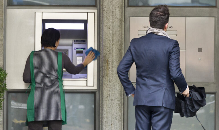 Deux personnes se tiennent devant la façade d'une banque. À gauche, une femme portant un gilet rayé vert et blanc nettoye un distributeur automatique de billets (DAB). À droite, un homme en costume bleu marine, tenant une serviette noire, attend devant la porte d'entrée de l'établissement bancaire.