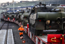 Loi sur le matériel de guerre : les enjeux de la votation de novembre Plusieurs chars de combat chargés sur des wagons ferroviaires sont stationnés sur une voie ferrée, tandis qu'un employé en tenue de sécurité orange marche le long du convoi. La scène montre le transport de véhicules militaires par train.