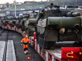 Loi sur le matériel de guerre : les enjeux de la votation de novembre Plusieurs chars de combat chargés sur des wagons ferroviaires sont stationnés sur une voie ferrée, tandis qu'un employé en tenue de sécurité orange marche le long du convoi. La scène montre le transport de véhicules militaires par train.