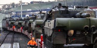 Loi sur le matériel de guerre : les enjeux de la votation de novembre Plusieurs chars de combat chargés sur des wagons ferroviaires sont stationnés sur une voie ferrée, tandis qu'un employé en tenue de sécurité orange marche le long du convoi. La scène montre le transport de véhicules militaires par train.