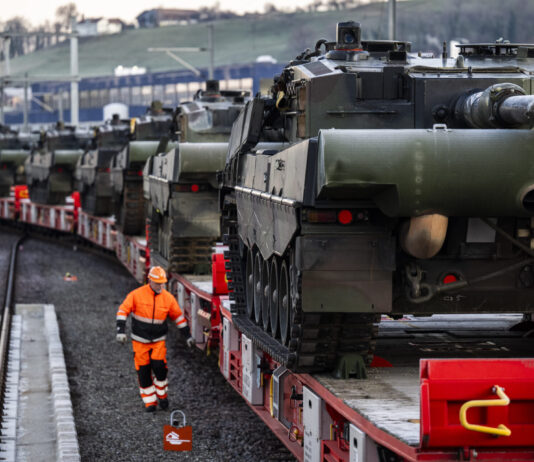 Loi sur le matériel de guerre : les enjeux de la votation de novembre Plusieurs chars de combat chargés sur des wagons ferroviaires sont stationnés sur une voie ferrée, tandis qu'un employé en tenue de sécurité orange marche le long du convoi. La scène montre le transport de véhicules militaires par train.