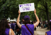L’affaire Frei le montre : la protection des victimes doit être renforcée "Une manifestante portant un ruban et un t-shirt violets tient une pancarte blanche avec le message 'VICTIMES on vous croit' écrit en lettres violettes, lors d'une marche de protestation dans un parc arborisé entouré d'autres participants."