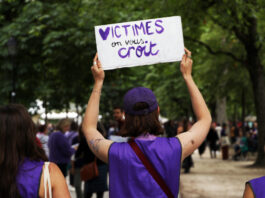 L’affaire Frei le montre : la protection des victimes doit être renforcée "Une manifestante portant un ruban et un t-shirt violets tient une pancarte blanche avec le message 'VICTIMES on vous croit' écrit en lettres violettes, lors d'une marche de protestation dans un parc arborisé entouré d'autres participants."