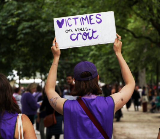 L’affaire Frei le montre : la protection des victimes doit être renforcée "Une manifestante portant un ruban et un t-shirt violets tient une pancarte blanche avec le message 'VICTIMES on vous croit' écrit en lettres violettes, lors d'une marche de protestation dans un parc arborisé entouré d'autres participants."