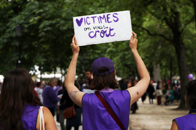 "Une manifestante portant un ruban et un t-shirt violets tient une pancarte blanche avec le message 'VICTIMES on vous croit' écrit en lettres violettes, lors d'une marche de protestation dans un parc arborisé entouré d'autres participants."