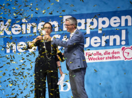 PLR : le prétendu parti de l’économie néglige sa comptabilité Deux personnes en tenues formelles célèbrent devant un grand panneau bleu avec du texte en allemand, tandis que des confettis dorés et noirs tombent autour d'elles. La femme à gauche porte des lunettes et un costume noir, l'homme à droite porte un costume bleu. Le panneau affiche "Keinem oppen" et "nern!" avec un logo rose et le texte "Für alle, die den Wecker stellen."
