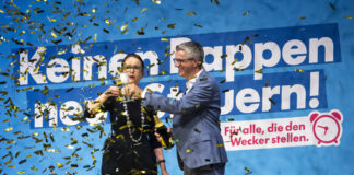 PLR : le prétendu parti de l’économie néglige sa comptabilité Deux personnes en tenues formelles célèbrent devant un grand panneau bleu avec du texte en allemand, tandis que des confettis dorés et noirs tombent autour d'elles. La femme à gauche porte des lunettes et un costume noir, l'homme à droite porte un costume bleu. Le panneau affiche "Keinem oppen" et "nern!" avec un logo rose et le texte "Für alle, die den Wecker stellen."