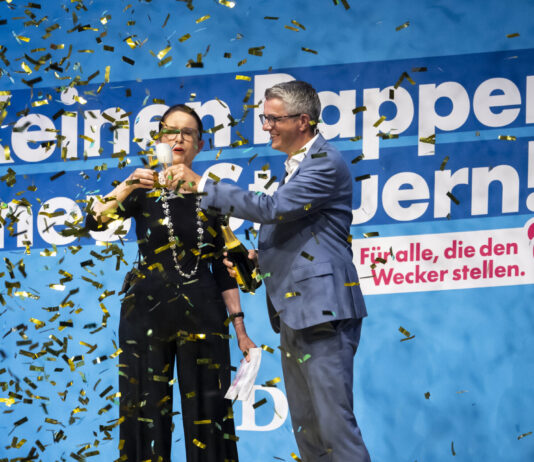 PLR : le prétendu parti de l’économie néglige sa comptabilité Deux personnes en tenues formelles célèbrent devant un grand panneau bleu avec du texte en allemand, tandis que des confettis dorés et noirs tombent autour d'elles. La femme à gauche porte des lunettes et un costume noir, l'homme à droite porte un costume bleu. Le panneau affiche "Keinem oppen" et "nern!" avec un logo rose et le texte "Für alle, die den Wecker stellen."