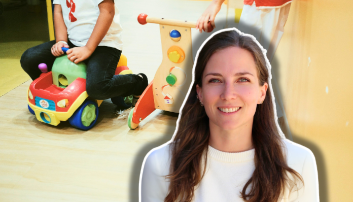 Femme aux cheveux longs, souriante à la caméra, vêtue d'un pull blanc crème, composée sur une image d'arrière-plan montrant un jeune enfant en train de jouer avec des jeux éducatifs colorés : un porteur jouet rouge et jaune avec des roues, et un jouet de frappe en bois avec des perles de couleurs.