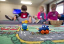 Voici comment la nouvelle loi sur les crèches améliorera la situation des familles Deux petites voitures jouets bleus et oranges sur un tapis de jeu routier coloré, avec quatre enfants assis en arrière-plan dans une salle de jeu lumineuse, jouant ensemble.