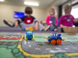 Voici comment la nouvelle loi sur les crèches améliorera la situation des familles Deux petites voitures jouets bleus et oranges sur un tapis de jeu routier coloré, avec quatre enfants assis en arrière-plan dans une salle de jeu lumineuse, jouant ensemble.