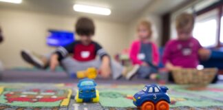 Voici comment la nouvelle loi sur les crèches améliorera la situation des familles Deux petites voitures jouets bleus et oranges sur un tapis de jeu routier coloré, avec quatre enfants assis en arrière-plan dans une salle de jeu lumineuse, jouant ensemble.