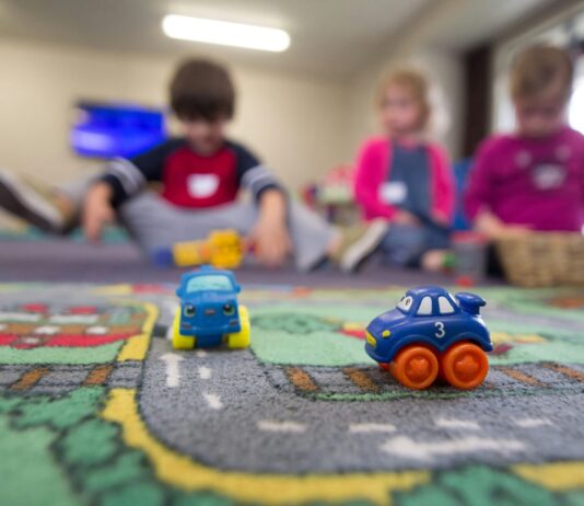 Voici comment la nouvelle loi sur les crèches améliorera la situation des familles Deux petites voitures jouets bleus et oranges sur un tapis de jeu routier coloré, avec quatre enfants assis en arrière-plan dans une salle de jeu lumineuse, jouant ensemble.