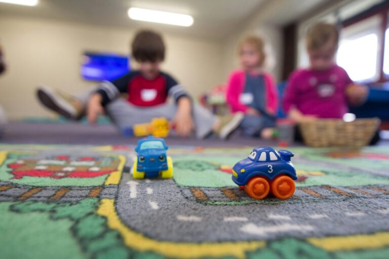 Deux petites voitures jouets bleus et oranges sur un tapis de jeu routier coloré, avec quatre enfants assis en arrière-plan dans une salle de jeu lumineuse, jouant ensemble.