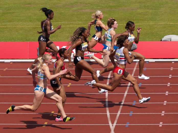 Huit sprinteuses disputent une course sur une piste d’athlétisme. Photographiées de profil en pleine accélération, elles courent dans leurs couloirs respectifs, à quelques mètres de la ligne d’arrivée, lors d’une compétition en plein air.