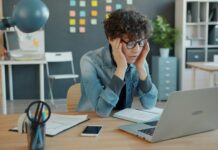 Réduction du temps de travail : la crainte des patrons face à des salarié-es en bonne santé Une personne aux cheveux bouclés portant des lunettes bleues et une chemise en denim est assise à un bureau en bois. Elle se tient la tête entre les mains dans une posture de réflexion ou de fatigue. Devant elle se trouvent un ordinateur portable, un smartphone, un bloc-notes et des fournitures de bureau (stylos, lampe bleue). L'arrière-plan montre un espace de travail moderne avec des rangements, des post-it colorés et des plantes décoratives.
