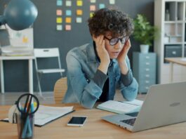 Réduction du temps de travail : la crainte des patrons face à des salarié-es en bonne santé Une personne aux cheveux bouclés portant des lunettes bleues et une chemise en denim est assise à un bureau en bois. Elle se tient la tête entre les mains dans une posture de réflexion ou de fatigue. Devant elle se trouvent un ordinateur portable, un smartphone, un bloc-notes et des fournitures de bureau (stylos, lampe bleue). L'arrière-plan montre un espace de travail moderne avec des rangements, des post-it colorés et des plantes décoratives.