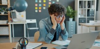 Réduction du temps de travail : la crainte des patrons face à des salarié-es en bonne santé Une personne aux cheveux bouclés portant des lunettes bleues et une chemise en denim est assise à un bureau en bois. Elle se tient la tête entre les mains dans une posture de réflexion ou de fatigue. Devant elle se trouvent un ordinateur portable, un smartphone, un bloc-notes et des fournitures de bureau (stylos, lampe bleue). L'arrière-plan montre un espace de travail moderne avec des rangements, des post-it colorés et des plantes décoratives.