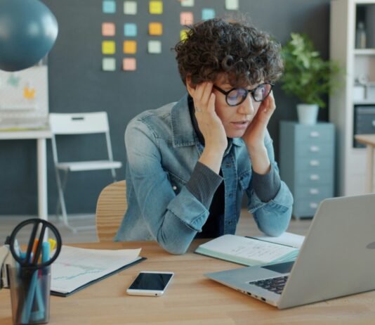 Réduction du temps de travail : la crainte des patrons face à des salarié-es en bonne santé Une personne aux cheveux bouclés portant des lunettes bleues et une chemise en denim est assise à un bureau en bois. Elle se tient la tête entre les mains dans une posture de réflexion ou de fatigue. Devant elle se trouvent un ordinateur portable, un smartphone, un bloc-notes et des fournitures de bureau (stylos, lampe bleue). L'arrière-plan montre un espace de travail moderne avec des rangements, des post-it colorés et des plantes décoratives.
