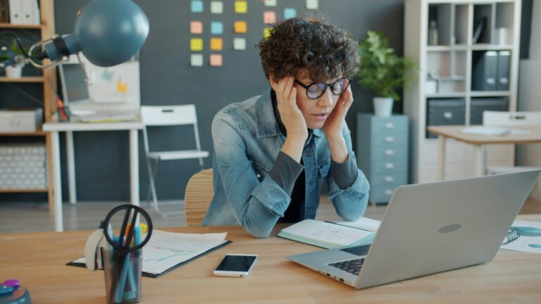 Une personne aux cheveux bouclés portant des lunettes bleues et une chemise en denim est assise à un bureau en bois. Elle se tient la tête entre les mains dans une posture de réflexion ou de fatigue. Devant elle se trouvent un ordinateur portable, un smartphone, un bloc-notes et des fournitures de bureau (stylos, lampe bleue). L'arrière-plan montre un espace de travail moderne avec des rangements, des post-it colorés et des plantes décoratives.