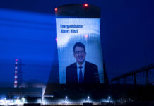 Albert Rösti supprime les mesures climatiques malgré les canicules records Projection nocturne sur une tour de refroidissement d'une centrale nucléaire suisse. L'écran géant affiche le portrait d'Albert Rösti, ministre suisse de l'énergie, accompagné du texte 'Energieminister Albert Rösti'. La tour, illuminée en bleu intense, est dotée de feux rouges clignotants au sommet. En arrière-plan, on distingue une cheminée striée rouge et blanc, tandis que le site industriel est éclairé par des projecteurs blancs au sol. Le ciel nocturne est d'un bleu profond.