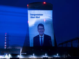 Albert Rösti supprime les mesures climatiques malgré les canicules extrêmes Projection nocturne sur une tour de refroidissement d'une centrale nucléaire suisse. L'écran géant affiche le portrait d'Albert Rösti, ministre suisse de l'énergie, accompagné du texte 'Energieminister Albert Rösti'. La tour, illuminée en bleu intense, est dotée de feux rouges clignotants au sommet. En arrière-plan, on distingue une cheminée striée rouge et blanc, tandis que le site industriel est éclairé par des projecteurs blancs au sol. Le ciel nocturne est d'un bleu profond.