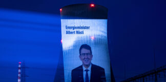 Albert Rösti supprime les mesures climatiques malgré les canicules extrêmes Projection nocturne sur une tour de refroidissement d'une centrale nucléaire suisse. L'écran géant affiche le portrait d'Albert Rösti, ministre suisse de l'énergie, accompagné du texte 'Energieminister Albert Rösti'. La tour, illuminée en bleu intense, est dotée de feux rouges clignotants au sommet. En arrière-plan, on distingue une cheminée striée rouge et blanc, tandis que le site industriel est éclairé par des projecteurs blancs au sol. Le ciel nocturne est d'un bleu profond.