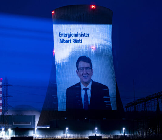 Albert Rösti supprime les mesures climatiques malgré les canicules records Projection nocturne sur une tour de refroidissement d'une centrale nucléaire suisse. L'écran géant affiche le portrait d'Albert Rösti, ministre suisse de l'énergie, accompagné du texte 'Energieminister Albert Rösti'. La tour, illuminée en bleu intense, est dotée de feux rouges clignotants au sommet. En arrière-plan, on distingue une cheminée striée rouge et blanc, tandis que le site industriel est éclairé par des projecteurs blancs au sol. Le ciel nocturne est d'un bleu profond.