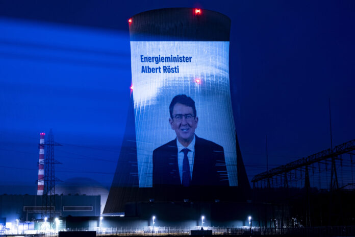 , AKW, ATOMKRAFT, AKTIVIST, GREENPEACE, PROJEKTION, AKW GOESGEN, ENERGIE, ATOMENERGIE, Projection nocturne sur une tour de refroidissement d'une centrale nucléaire suisse. L'écran géant affiche le portrait d'Albert Rösti, ministre suisse de l'énergie, accompagné du texte 'Energieminister Albert Rösti'. La tour, illuminée en bleu intense, est dotée de feux rouges clignotants au sommet. En arrière-plan, on distingue une cheminée striée rouge et blanc, tandis que le site industriel est éclairé par des projecteurs blancs au sol. Le ciel nocturne est d'un bleu profond.