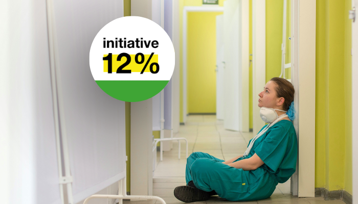 Une professionnelle de santé en uniforme vert menthe, assise dans un couloir d'établissement médical aux murs jaunes et blancs, regardant vers le haut un badge circulaire blanc et vert portant le texte "initiative 12%" en noir et jaune.